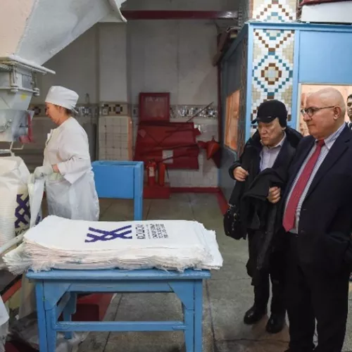 In a factory, workers in white uniforms handle sacks at a machine while people in business attire observe. Stacks of printed white sacks are on a blue table nearby.